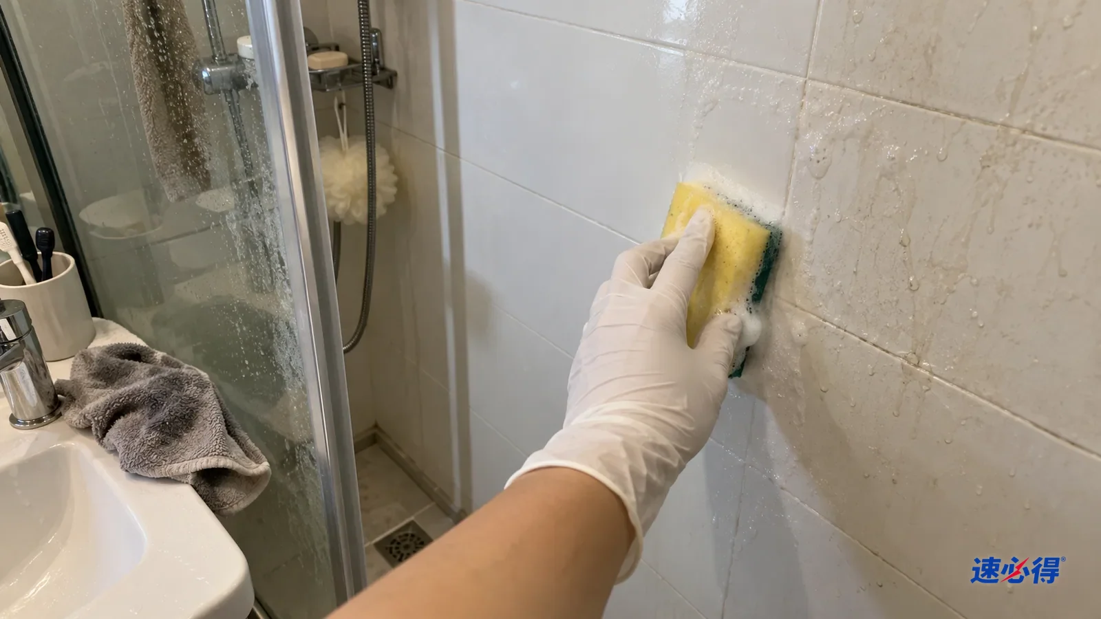 一只戴白色丁腈手套的手用软海绵擦淋浴区白瓷砖墙，海绵带着少量泡沫，擦过一块干净，旁边还有水渍，台面上盖着毛巾。