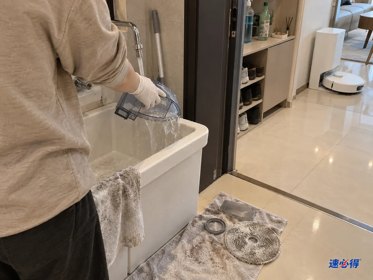 家人在一步外拍的半身背影，戴白色丁腈手套的手在玄关旁的拖把池里冲洗扫地机器人清水箱，水流从箱口溢出，旁边浅色瓷砖地面上铺着一块毛巾，上面放着拆下的密封圈和拖布。