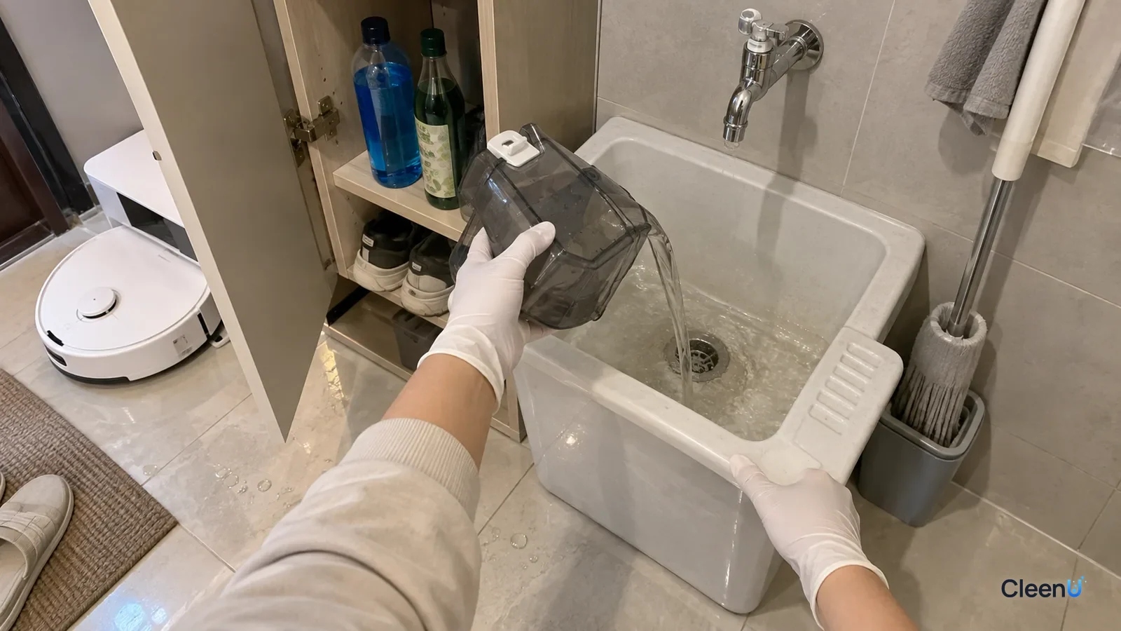 A gloved hand empties a robot mop clean water tank into the utility sink, glass cleaner and other bottles in the cabinet.