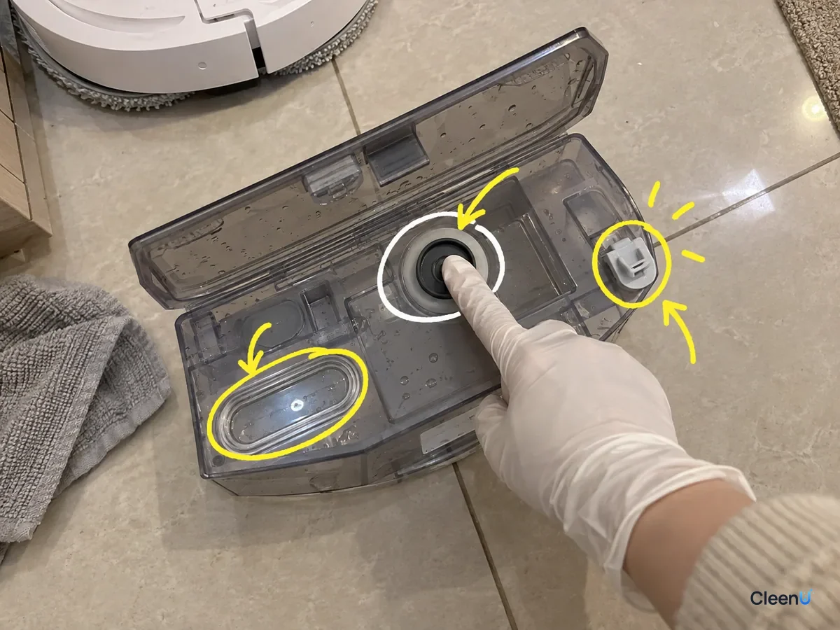 An open robot mop clean water tank with its outlet seal, level window and clip marked, a gloved finger pressing the seal.