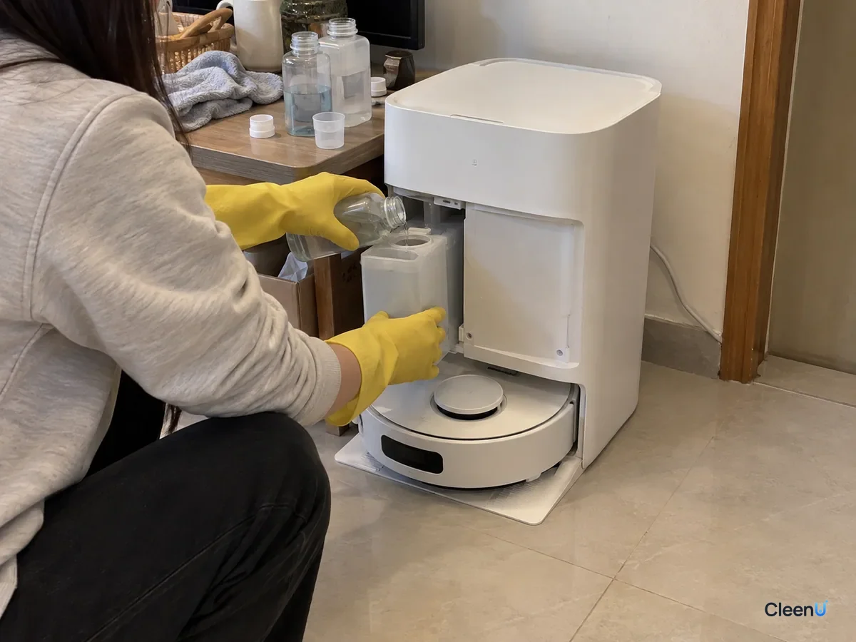 A person in yellow gloves crouches and slides a refilled solution cartridge into the side of a robot mop's auto-dosing dock.