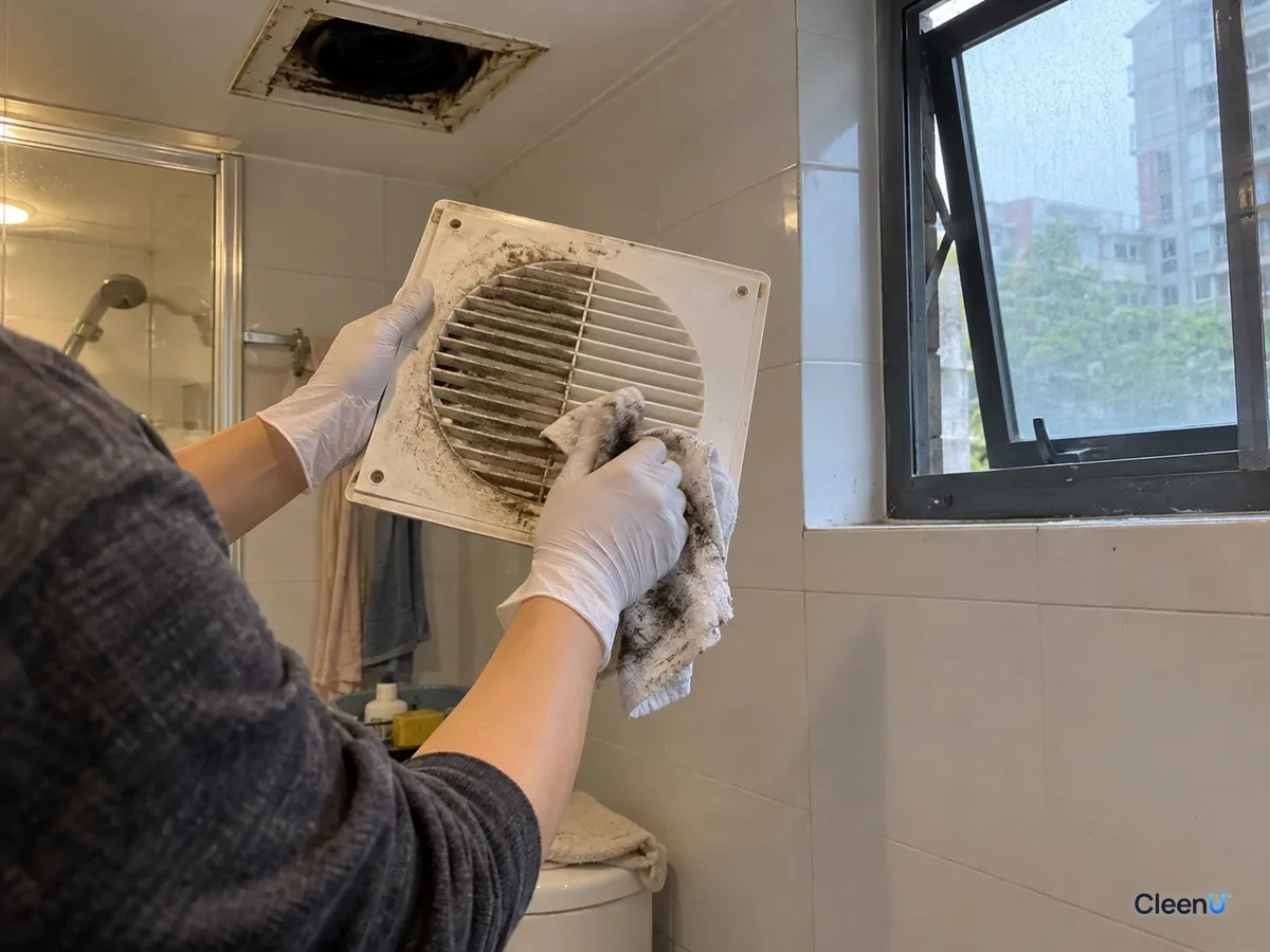 From a step back: a white nitrile-gloved hand wipes a removed exhaust fan cover with a damp cloth now gray-black with dust, half the cover clean, the bathroom window open.