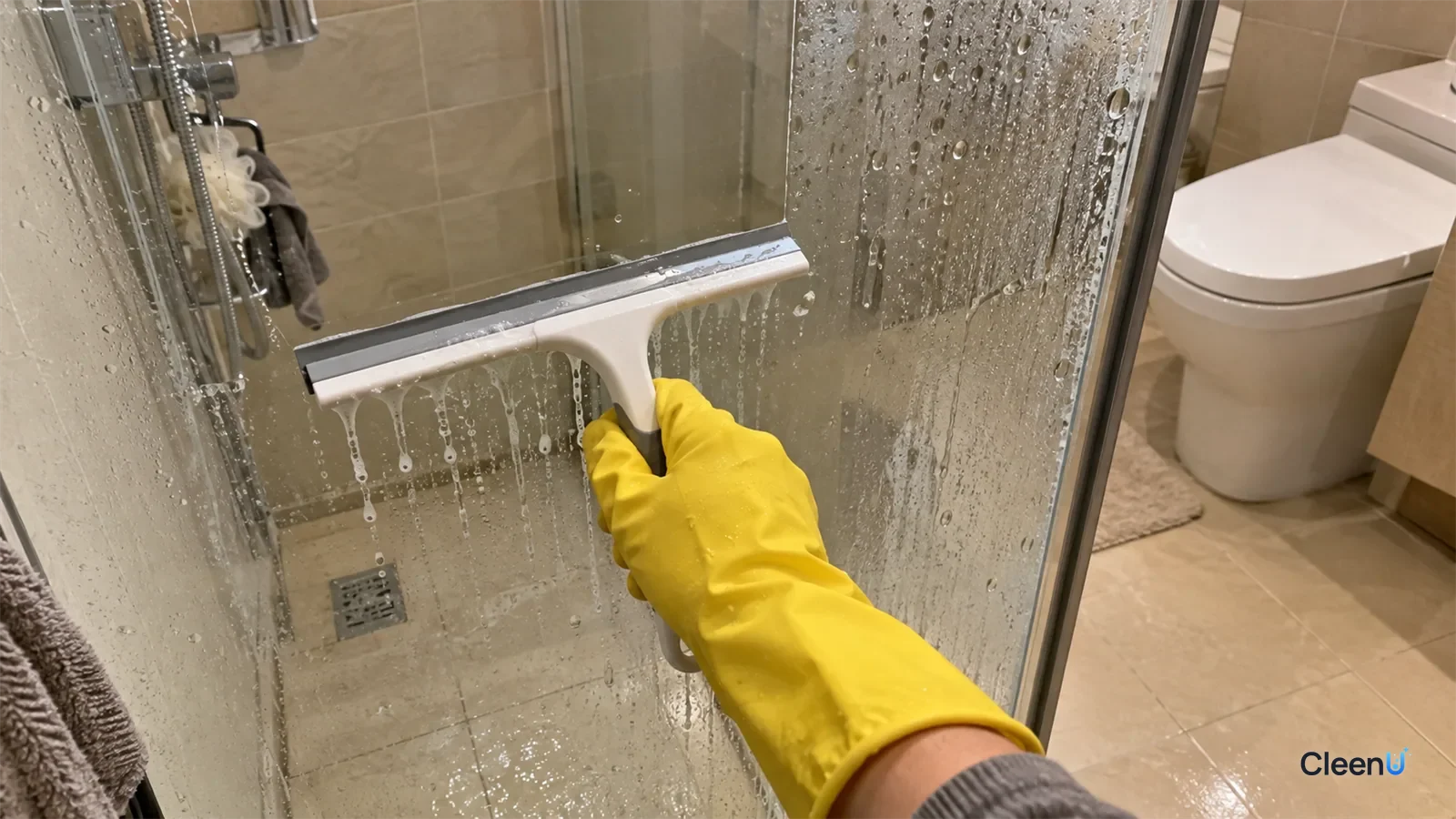 First-person: a yellow rubber-gloved hand squeegees the glass shower screen, one clear stripe with grey water running off the blade, water spots beside it.