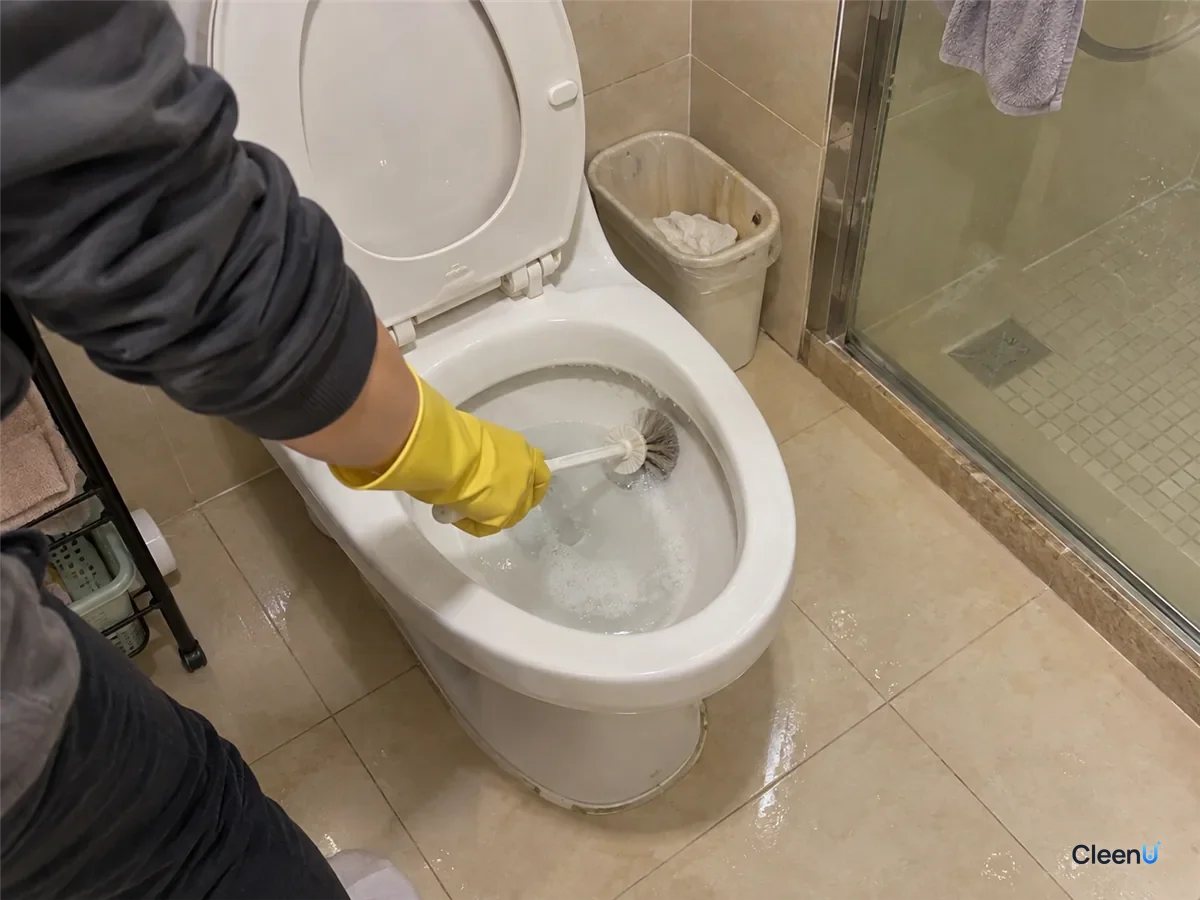 A yellow rubber-gloved hand scrubs under the toilet rim with a toilet brush, a cleaned stretch of white glaze showing and a little foam on the water.