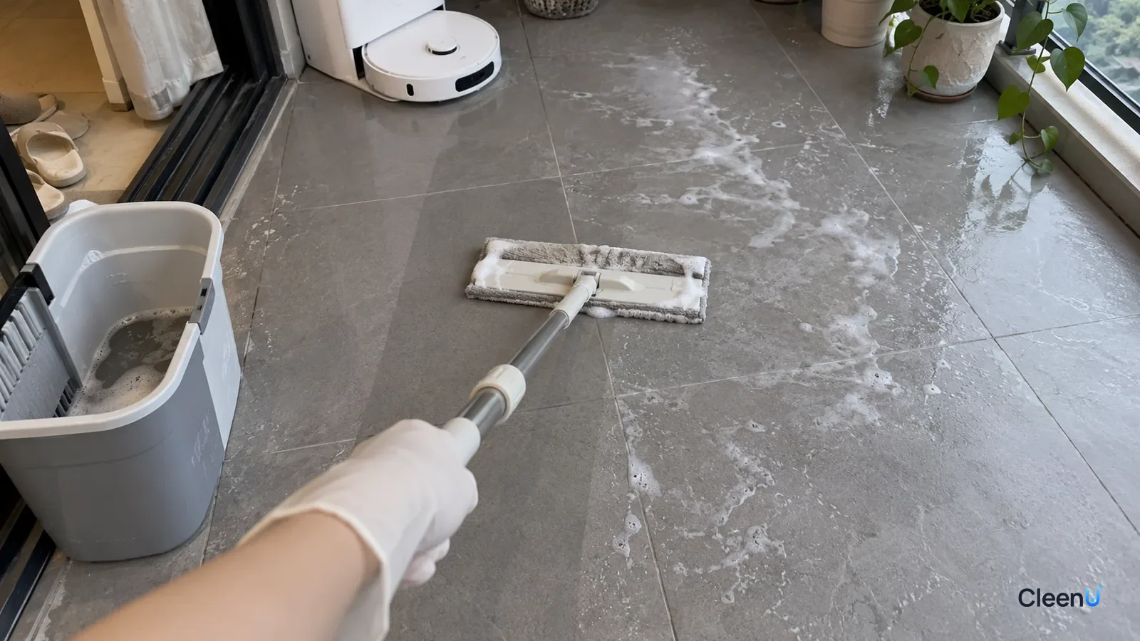 First-person close-up: a hand in white nitrile gloves pushes a rinsed flat mop over white soap streaks on gray matte balcony tile; a little foam on the pad, one strip clean, streaks beside it.