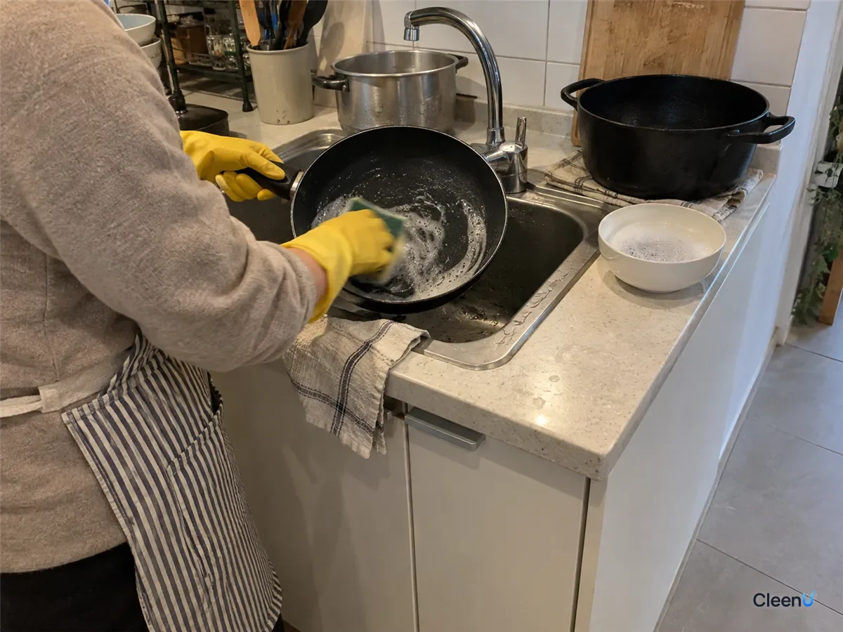 At the sink, yellow rubber-gloved hand washing a nonstick pan with a soft sponge and dish soap, a little grey foam in the pan, the dish soap bowl and cast iron pan beside
