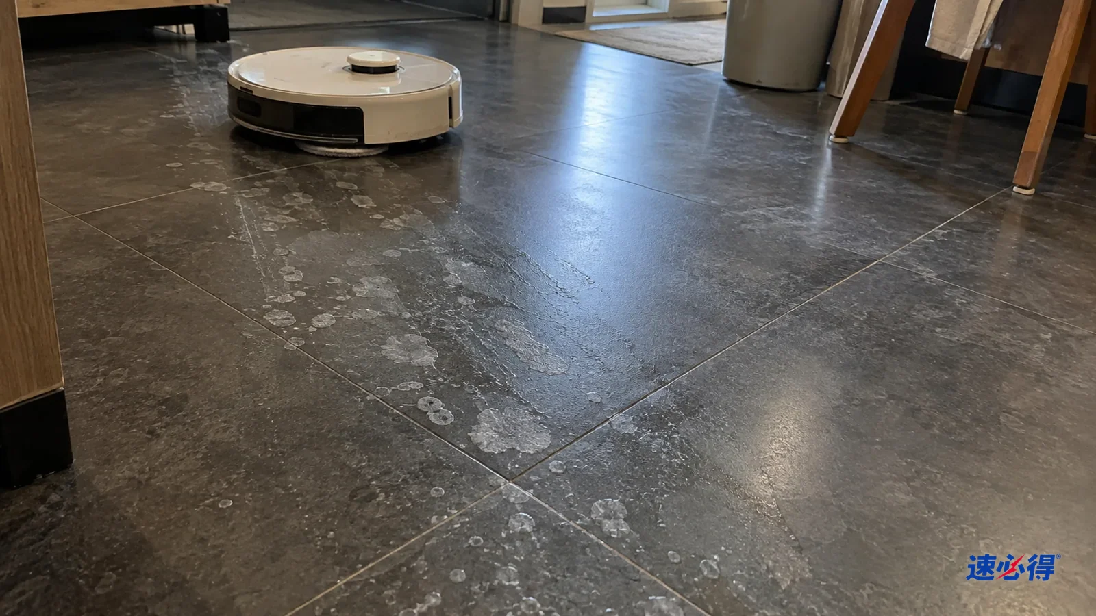 AI 示意图：深色瓷砖地面在侧光下，扫地机器人拖过的路径上留下一片片白色的水垢斑点