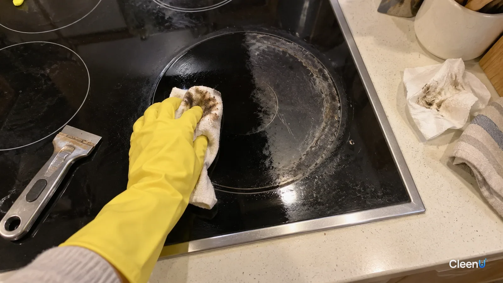 First-person close-up: a hand in yellow rubber gloves circles a soft cloth over a burnt ring on a black glass-ceramic cooktop; brown marks on the cloth, half clean, half still hazy.