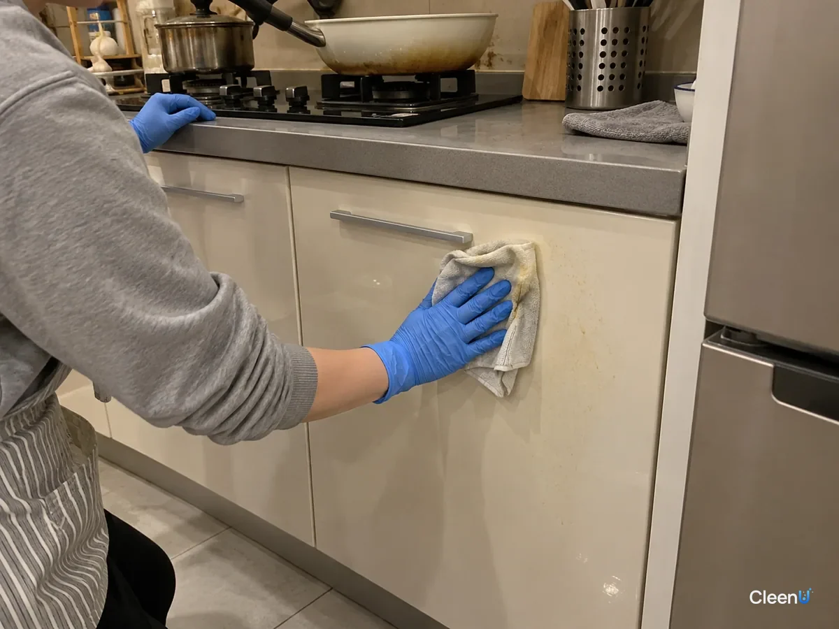 A blue nitrile-gloved hand wipes grease film off a light gloss cabinet door beside the stove with a damp microfiber cloth tinged yellow, leaving the aluminum bar handle untouched.