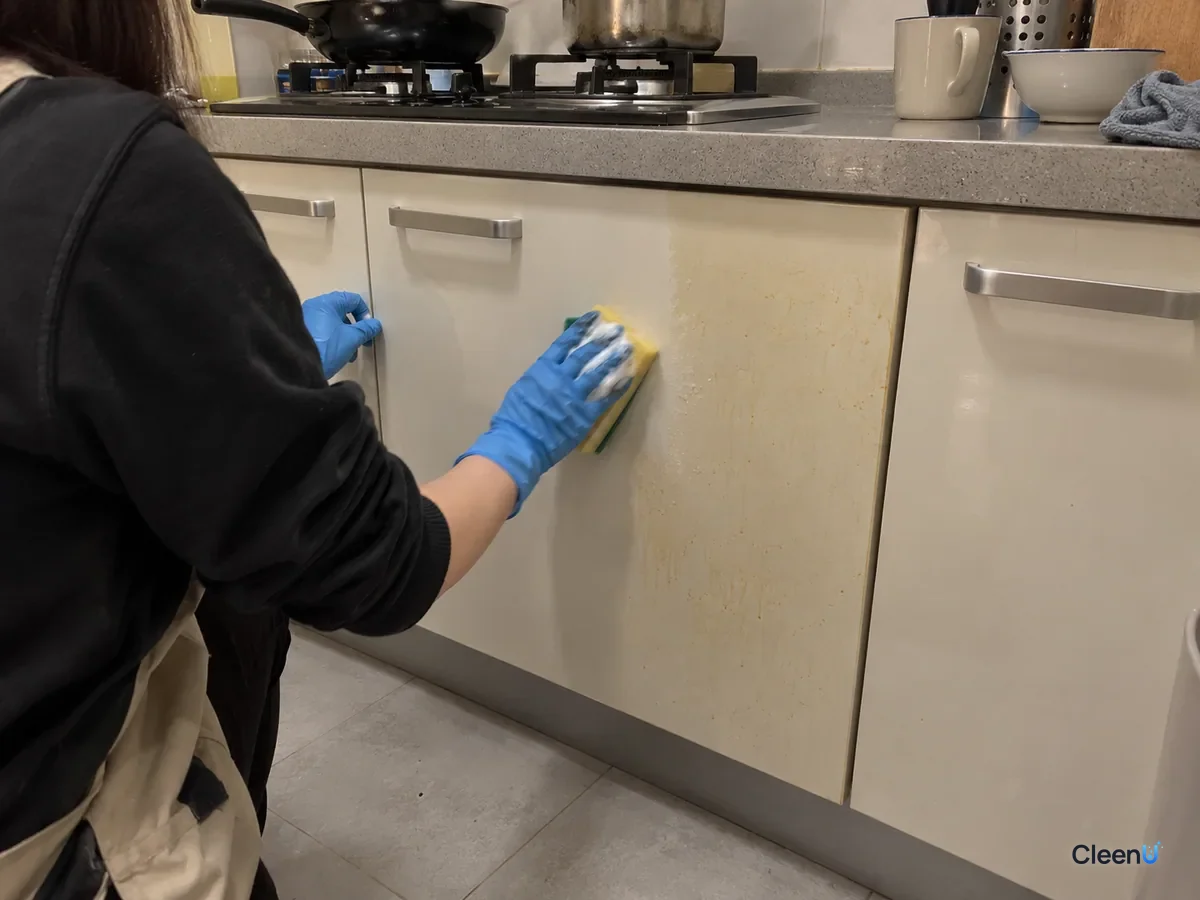 Half-body from behind: a blue nitrile-gloved hand sponges a light cabinet door by the stove, a little foam on the sponge, the wiped band clean beside still-yellowed panel.