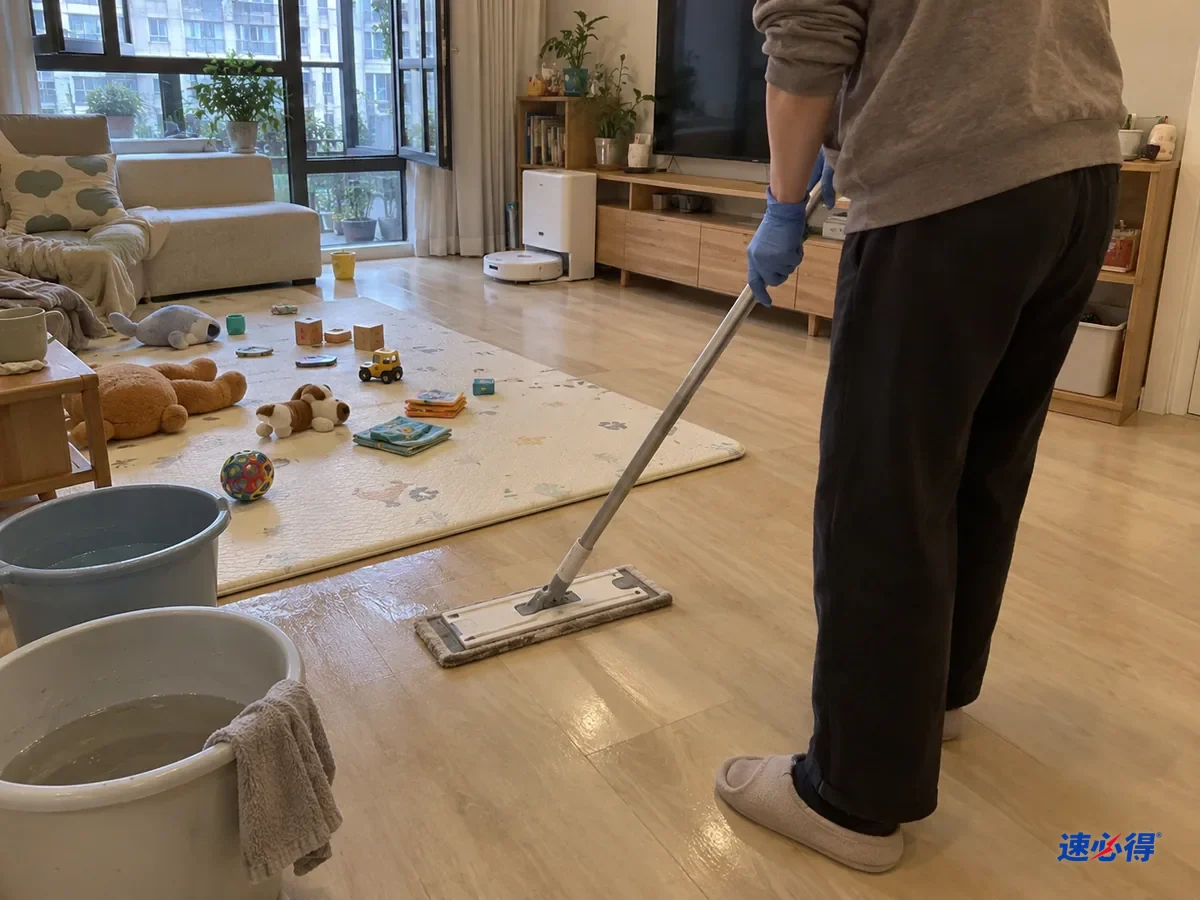 半身背影，戴蓝色丁腈手套的手推着平板拖把用清水再过一遍爬行垫周围地面，拖布边沾着少量灰，旁边两只水桶，窗户开着。