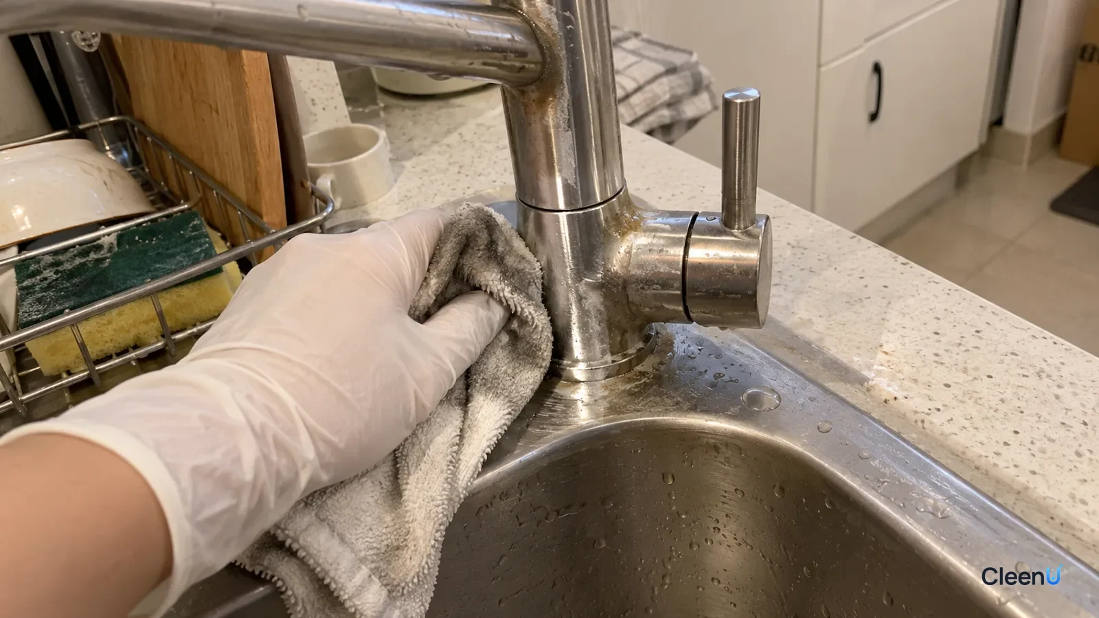 First-person: a white nitrile-gloved hand wipes grime off a stainless faucet base with a cloth, half shiny, grey dirt on the cloth.