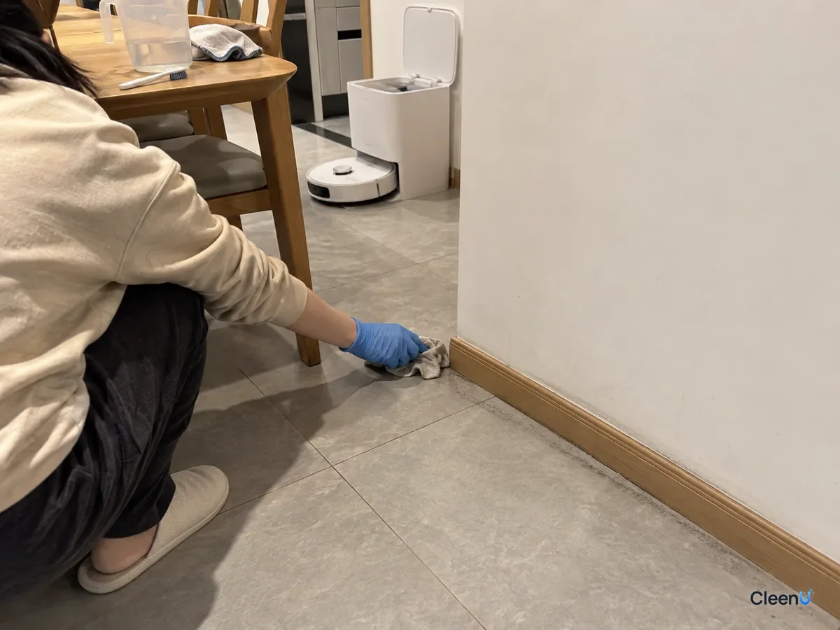 A blue-gloved hand wipes gray floor tile along the dining room baseboard with a damp cloth, the robot dock in the background.
