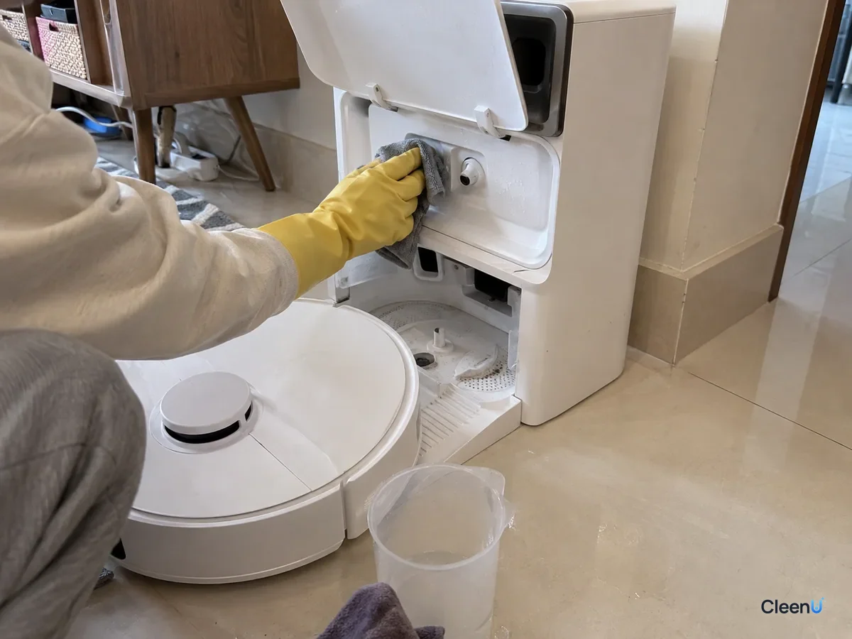 A yellow-gloved hand wipes drips from the dock's compartment outlet with a damp cloth, the robot vacuum parked below.