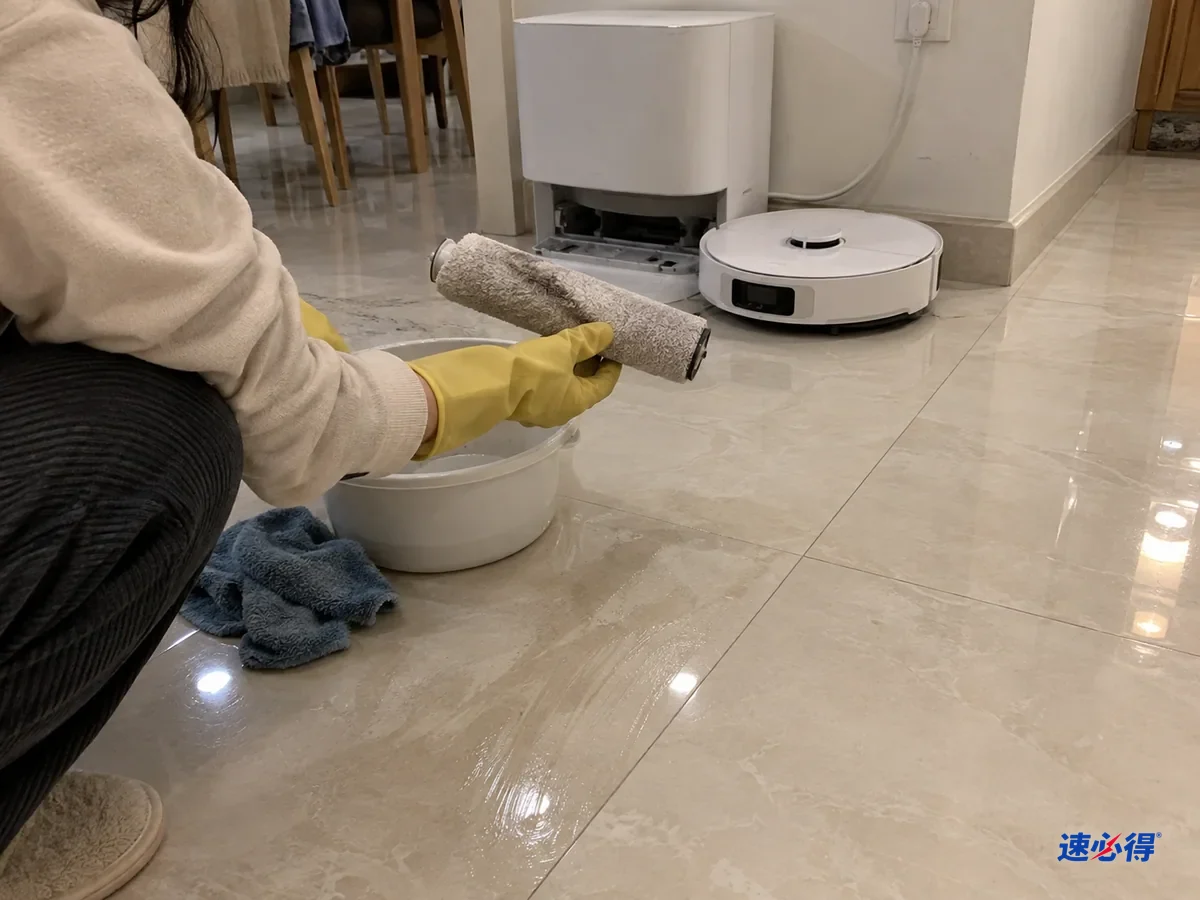 戴黄色手套的人蹲在基站前，拿起刚取出的滚筒拖布查看，地砖上留着未干拖痕