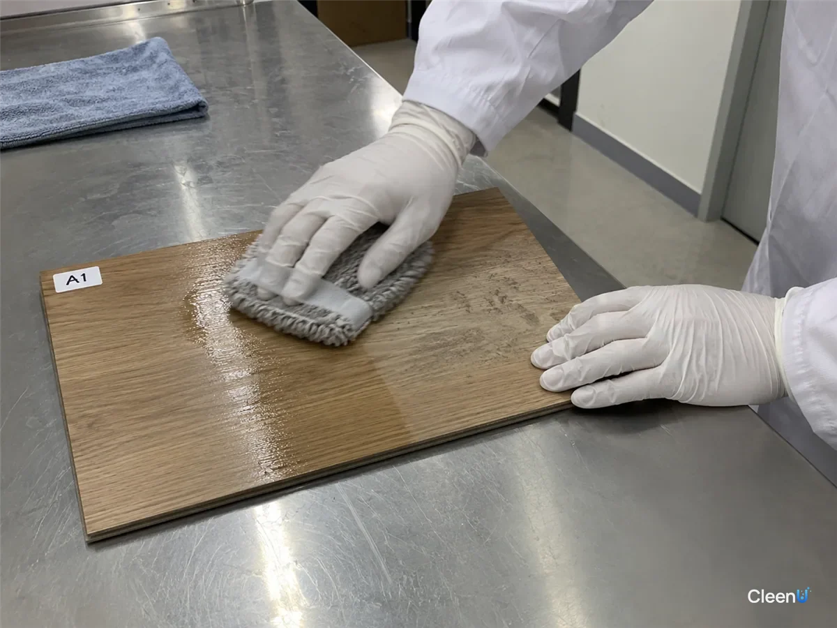 In the lab, a white nitrile-gloved hand and forearm push a mop pad across a laminate sample, a wet unfinished streak on one half, dusty marks on the other.