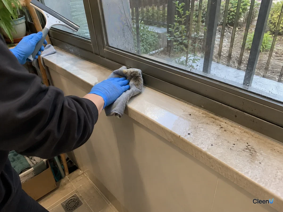Blue nitrile-gloved hand wiping the aluminum frame and stone sill with microfiber, muddy grey on the cloth, one stretch of sill clean, mud specks still on the rest