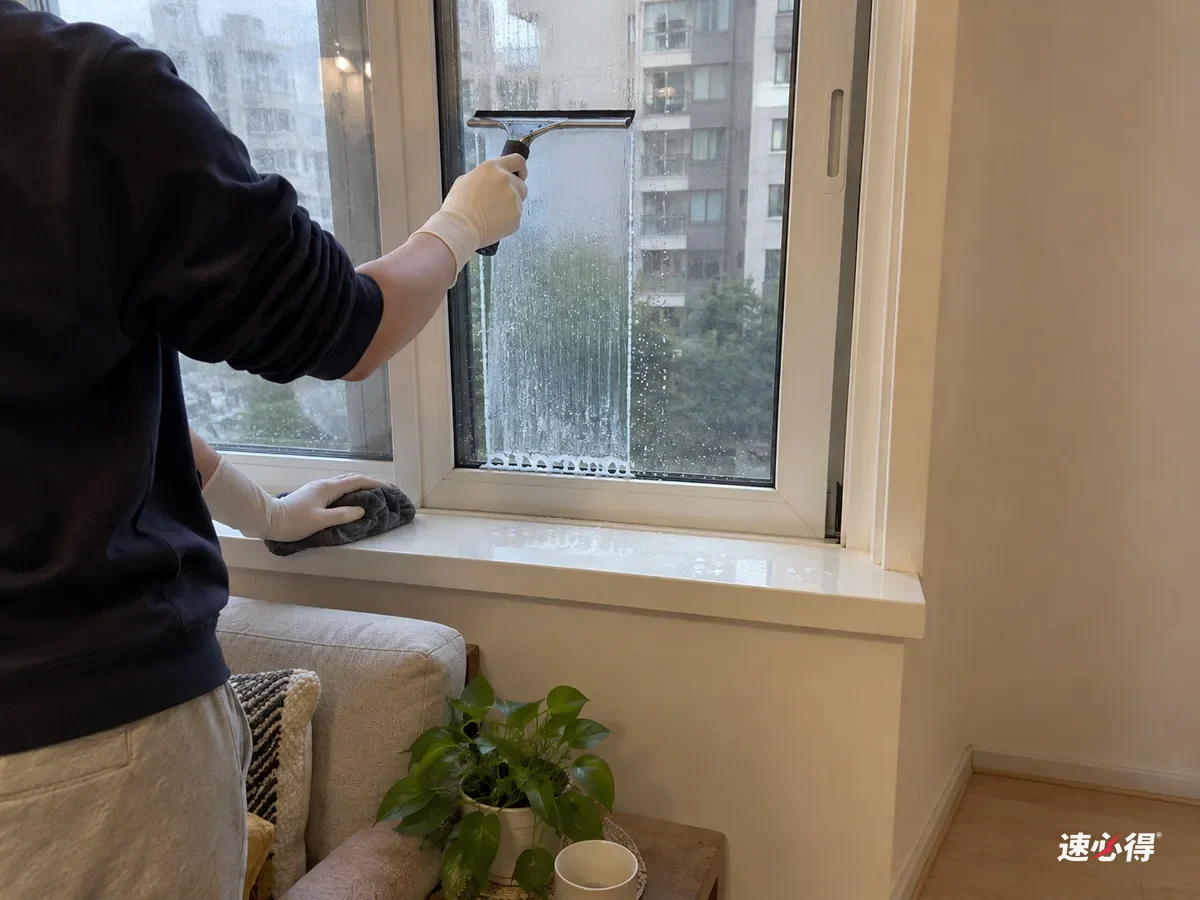 一步外看，戴白色丁腈手套的手拿刮水器从窗玻璃顶部往下刮，刮过的一条清楚，下沿积着一小串灰色水，白色窗台上滴着水点。
