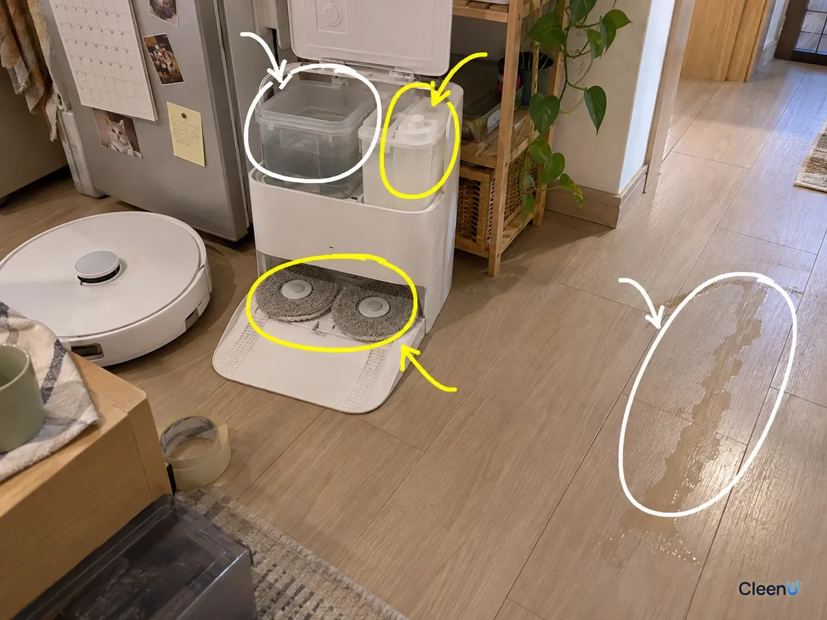 A robot mop dock with its clean water tank, solution bottle and mop pads marked, plus a damp streak left on the wood floor.