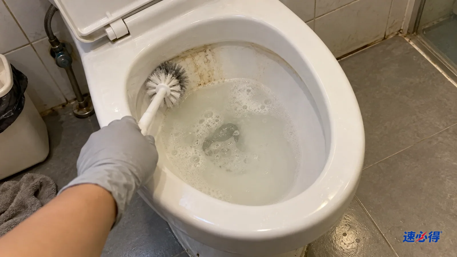 第一人称近拍：戴浅灰色丁腈手套的手握马桶刷刷马桶边缘下方的黄色印迹，刷过的一段变白，水面浮着少量泡沫。