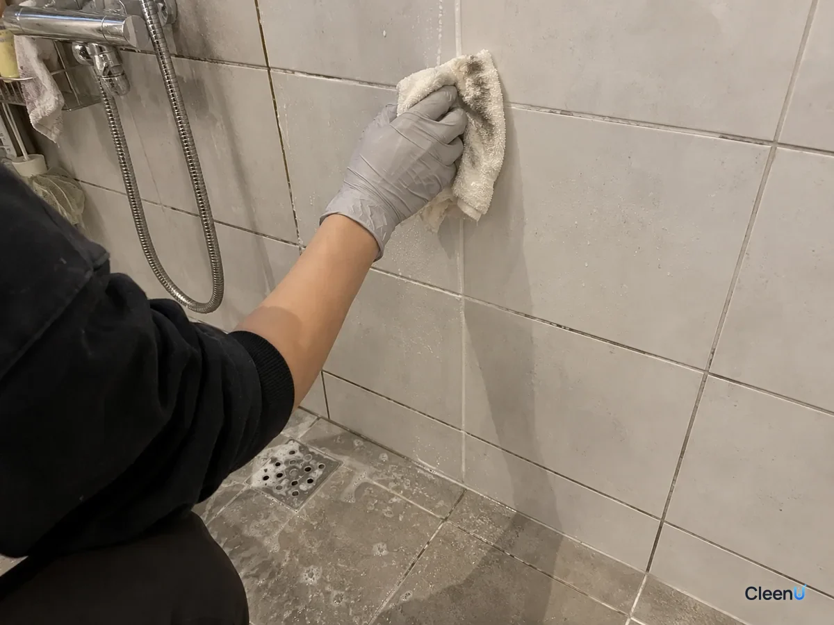 Same wall later: after rinsing, a hand in light grey nitrile gloves presses a cloth along the scrubbed grout to blot it dry; the cloth shows faint grey marks, the grout visibly lighter.