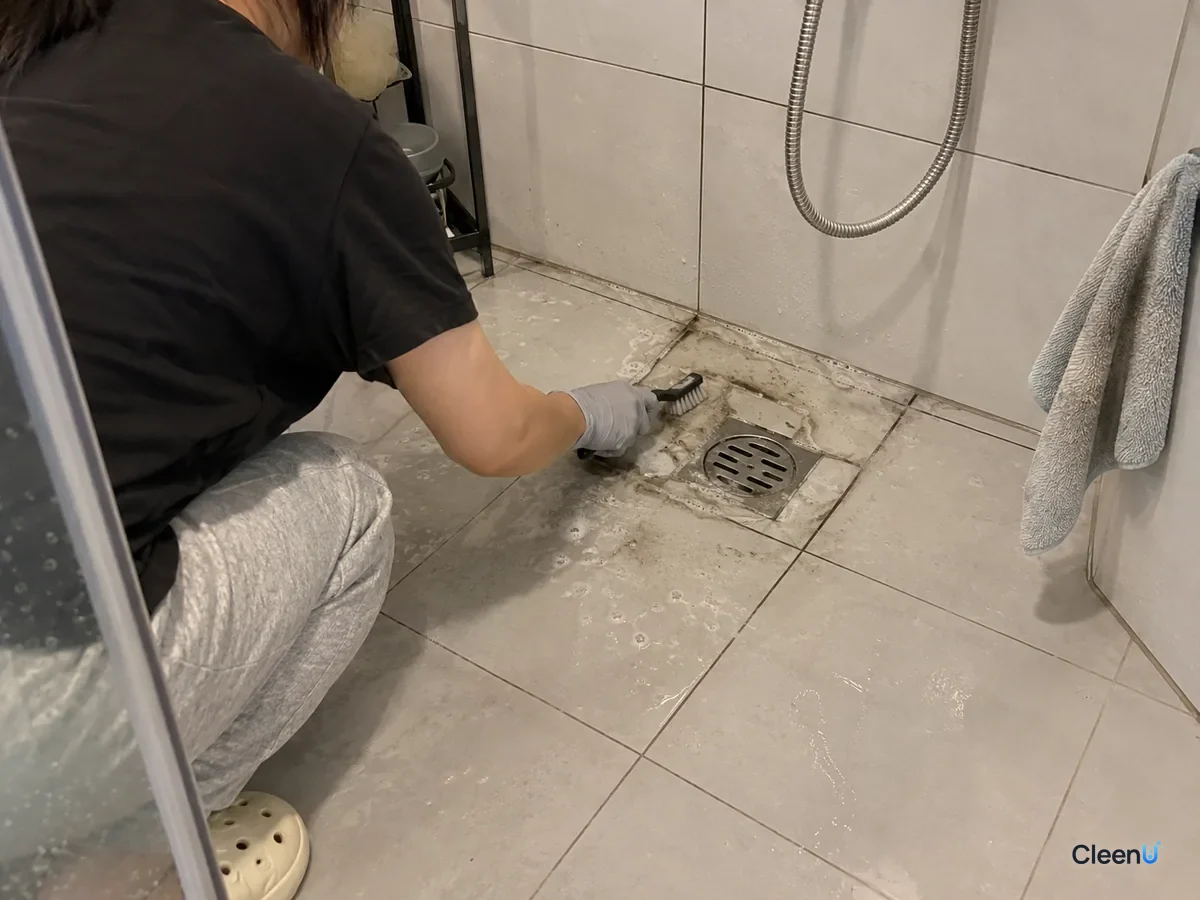 Seen from a step away: a crouching person, back and forearm only, in light gray nitrile gloves brushes floor grout by the drain; gray-black foam on the brush, a cloth on the wall ledge.