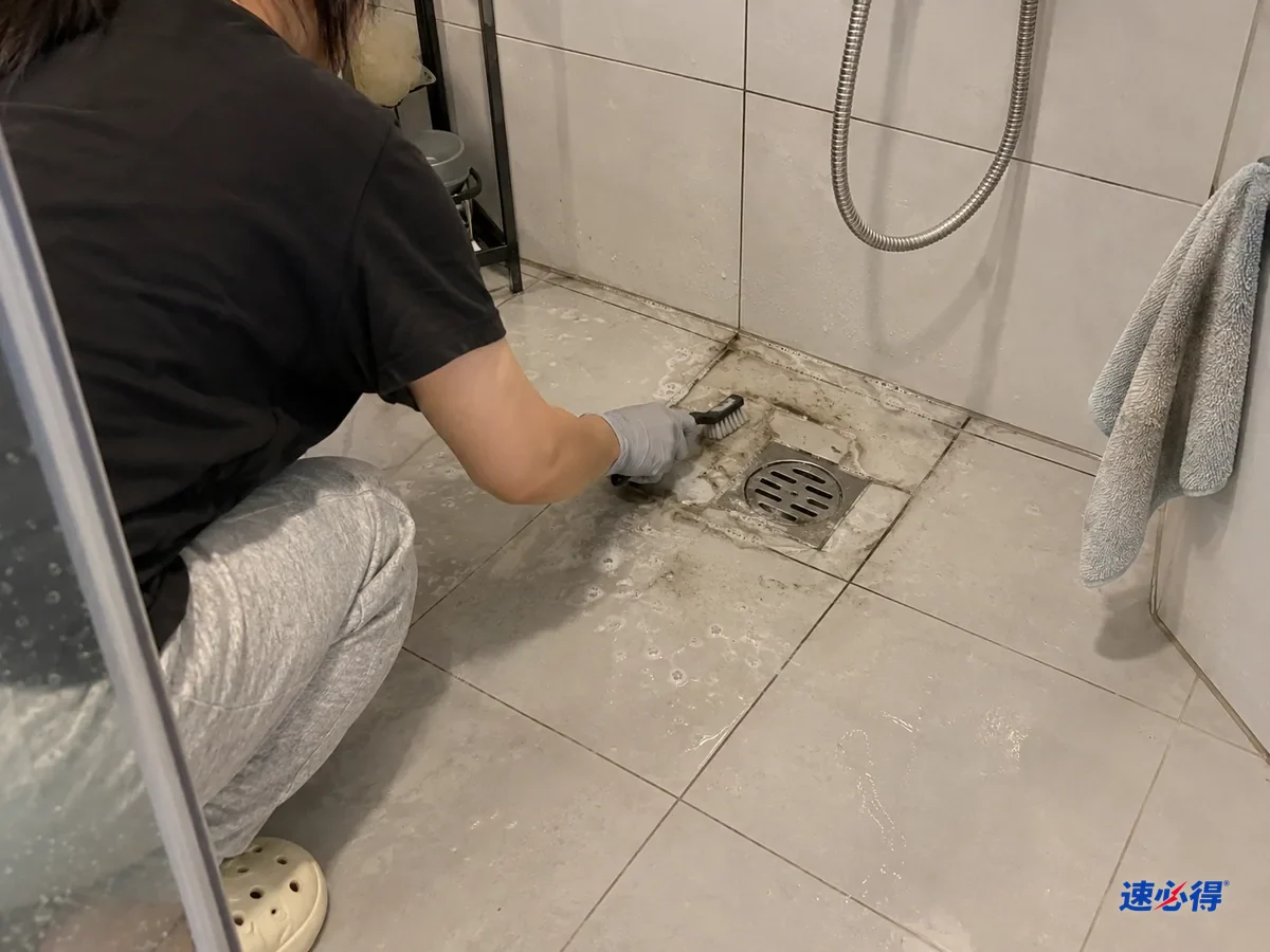 一两步外看，蹲在淋浴区的人的前臂和背影，戴浅灰色丁腈手套用窄头硬毛刷刷地漏边的地砖缝，刷头带着灰黑泡沫，吸水抹布搭在墙边。