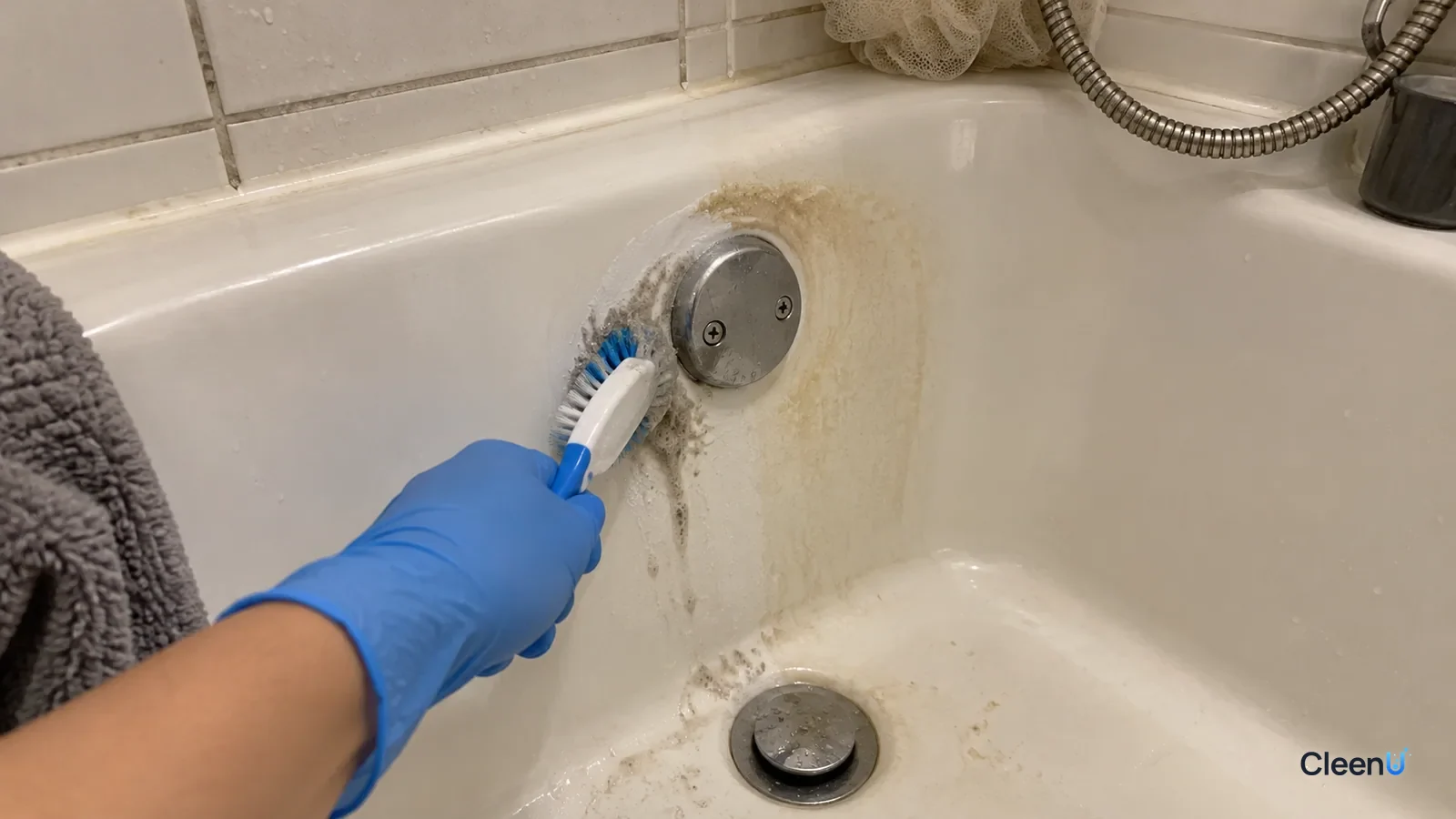 Blue nitrile-gloved hand brushing the greyed area around a tub drain, grey foam on the bristles, one ring white again, the faint yellow scum ring still on the wall