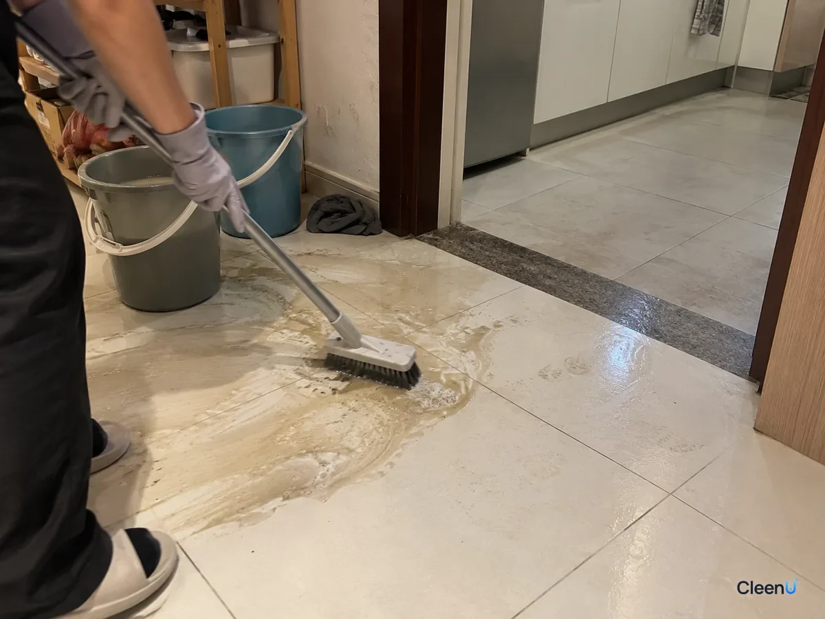 At the kitchen doorway a light gray nitrile-gloved hand scrubs the tile with a floor brush, raising gray-yellow murky water and a little foam beside still-dingy tiles.