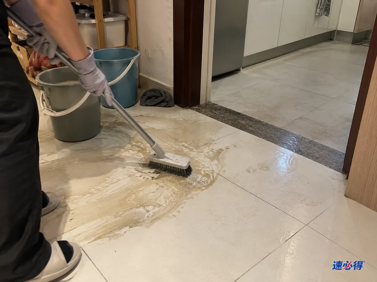 厨房门口，戴浅灰色丁腈手套的手握着地刷来回刷瓷砖地面，砖面上冒起一层灰黄色浑水和少量泡沫，旁边还没刷的砖发灰。
