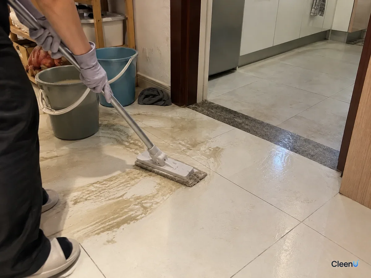 Same doorway: the gray-gloved hand mops up the loosened gray-yellow water while the floor is still wet; the mop pad has turned gray and the nearby bucket holds murky rinse water.