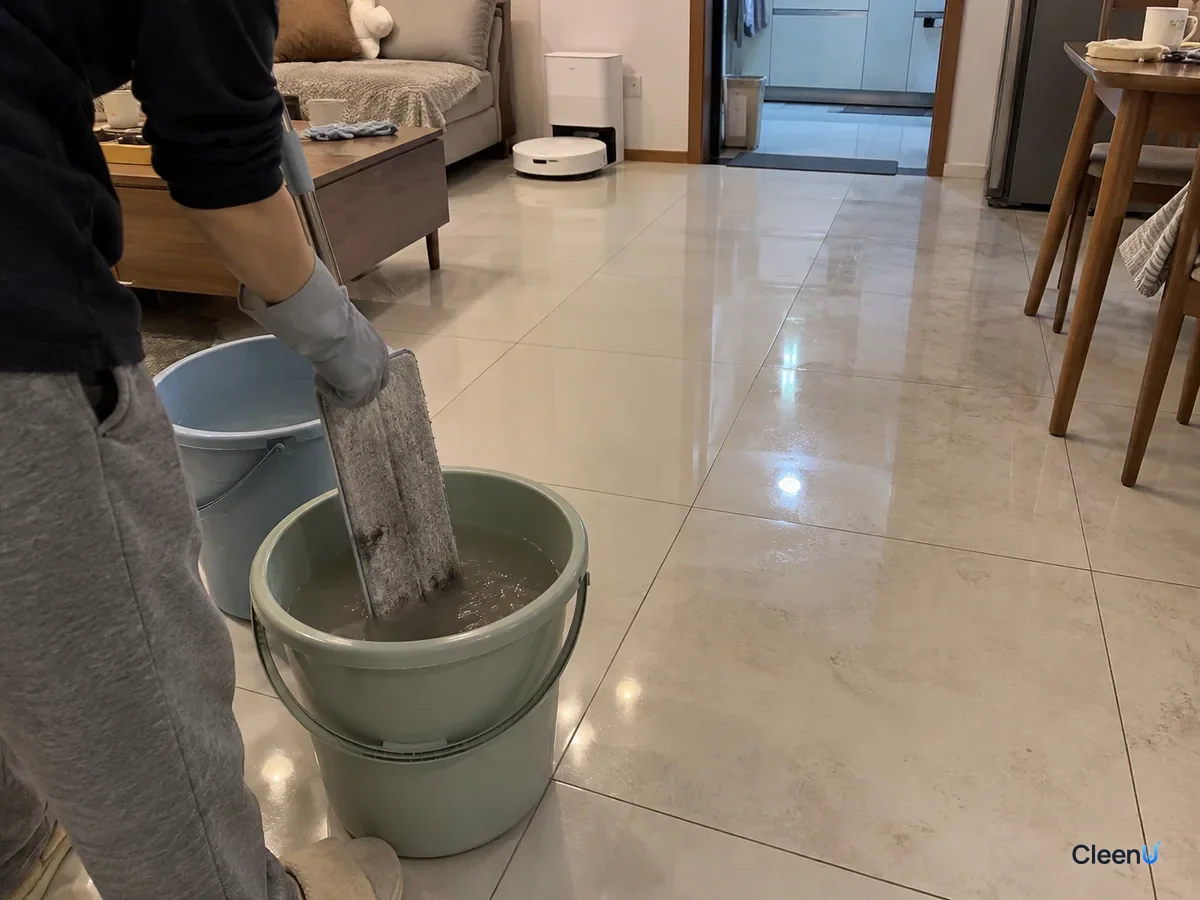 From a step back: a light gray nitrile-gloved hand rinses the grayed flat mop pad in the second bucket, water turning murky, the tile behind half done and half still dingy.