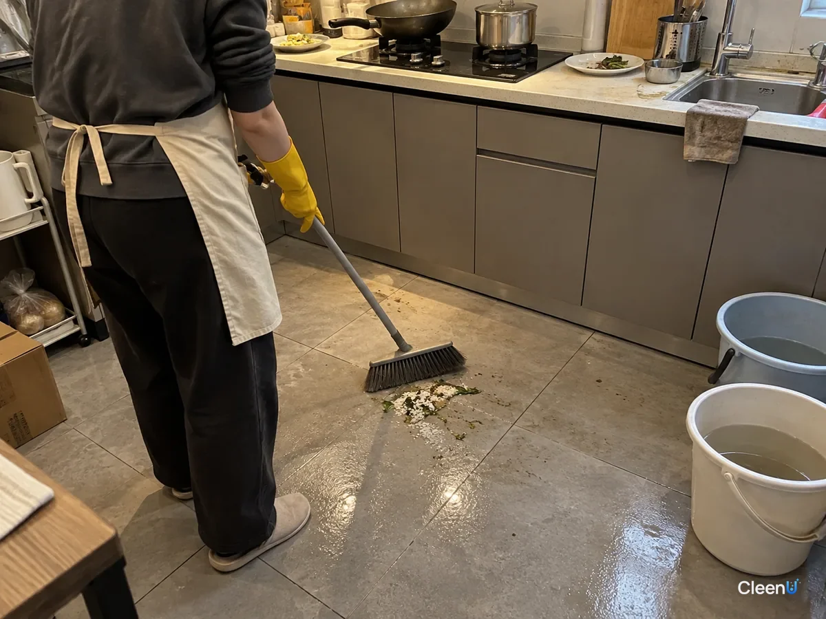 Person from behind in yellow rubber gloves sweeping rice grains, vegetable bits and grit into a small pile in front of the stove, the tiles still with a greasy sheen