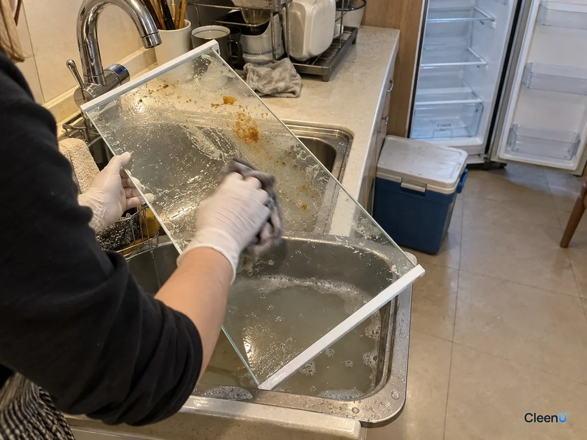 In the kitchen sink, white nitrile-gloved hand wiping a removed fridge glass shelf with a wet cloth, half still marked with soup rings, a little grey water in the sink