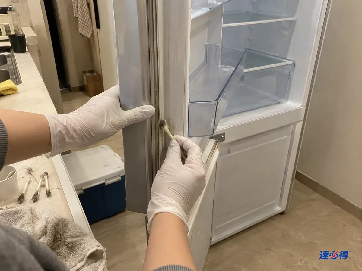戴白色丁腈手套的手用棉签沿冰箱门封条褶皱擦拭，棉签头沾上灰黑污垢，旁边放着几根用过的棉签