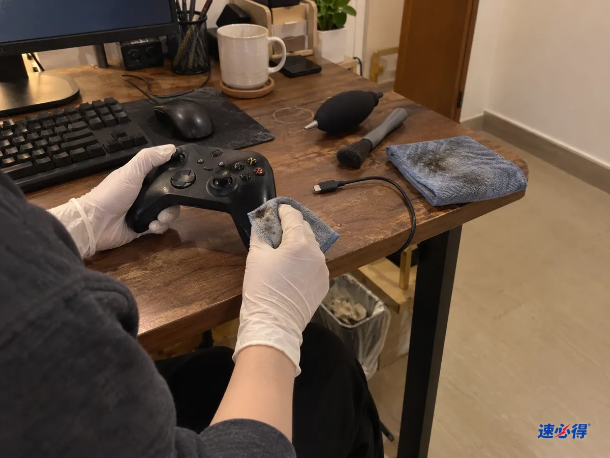 书桌上，戴白色丁腈手套的手用微湿的布擦游戏手柄的握把，布避开了旁边的充电口和按键缝隙，拔下的充电线放在一边。