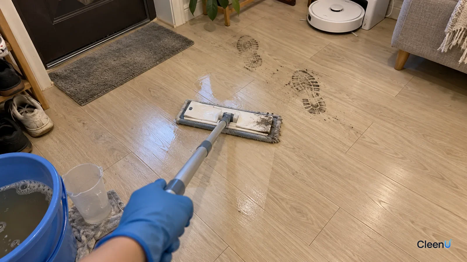 First-person close-up: a hand in blue nitrile gloves pushes a flat mop over gray shoe prints on light wood-look SPC planks by the door; gray dirt on the pad edge, two prints still ahead.