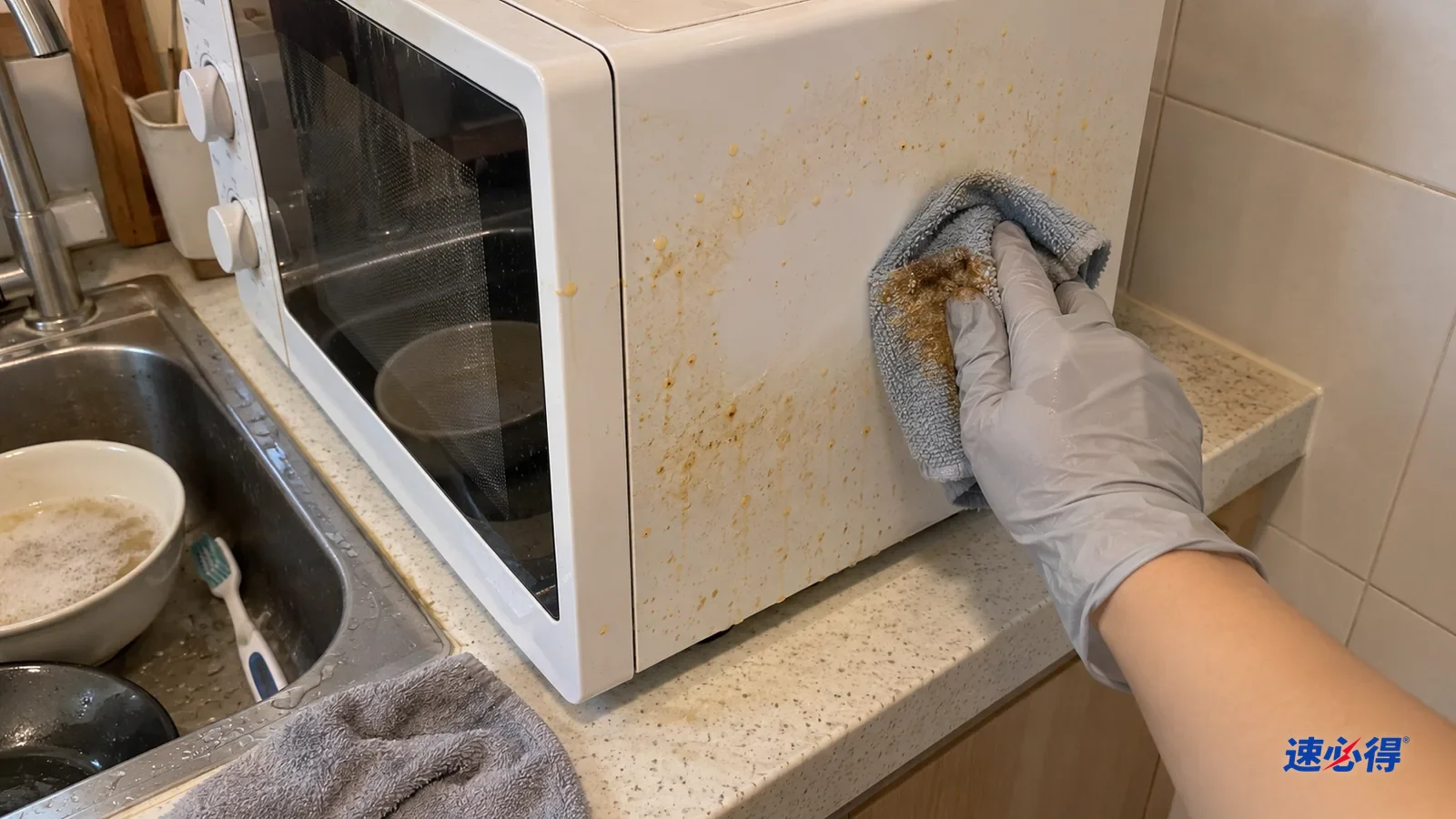 一只戴浅灰色丁腈手套的手用湿超细纤维布擦微波炉侧壁黄色油点，布上沾黄褐油迹，擦过一块变白，旁边还有油点。