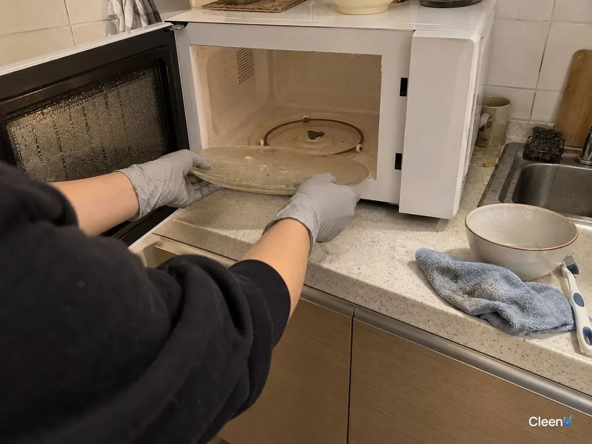Light grey nitrile-gloved hands lifting the glass turntable out of the microwave, steam haze still on the door glass, the roller ring and a few grease spots showing underneath