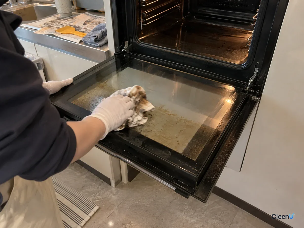 Same oven door later: the film scraped off, a hand in white nitrile gloves wipes the inner glass with a wrung damp cloth that shows brown grease, most of the glass now clear.