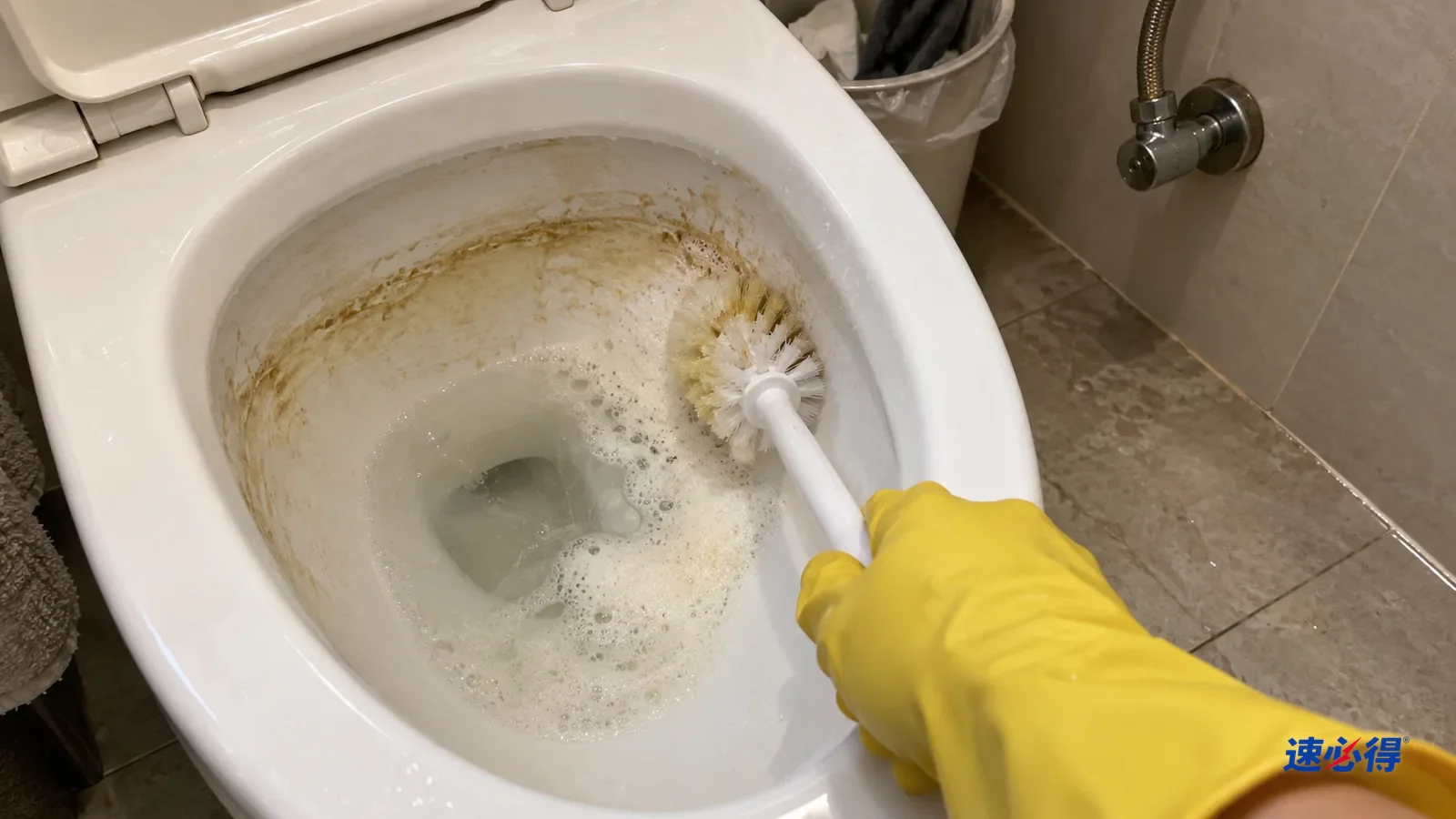 第一人称近拍，戴黄色橡胶手套的手用马桶刷刷白色马桶内壁水位线垢圈，刷毛带着淡黄泡沫，刷过一段露出白瓷，旁边仍是黄褐色垢圈。