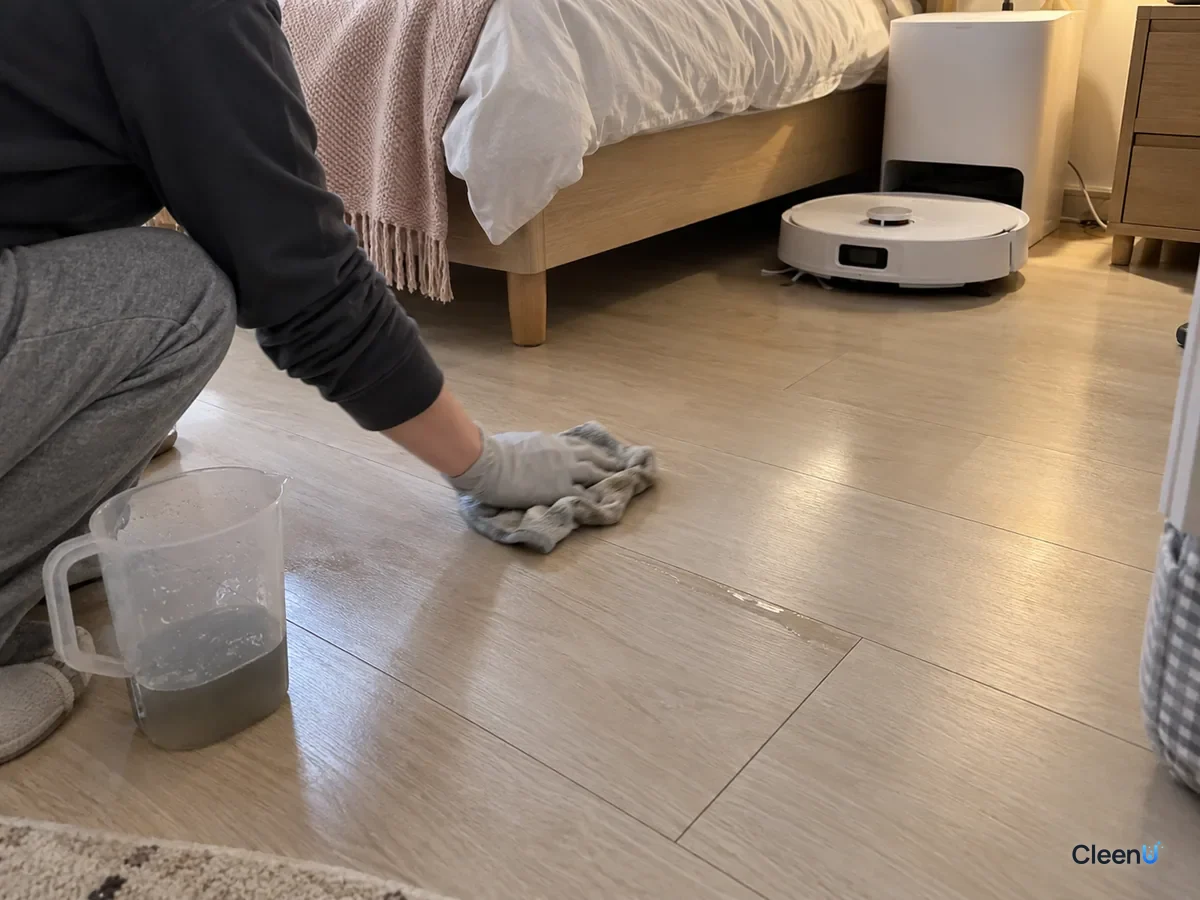A gloved hand wipes a laminate floor plank with a damp cloth, a jug of gray water beside it and the robot mop by the bed.