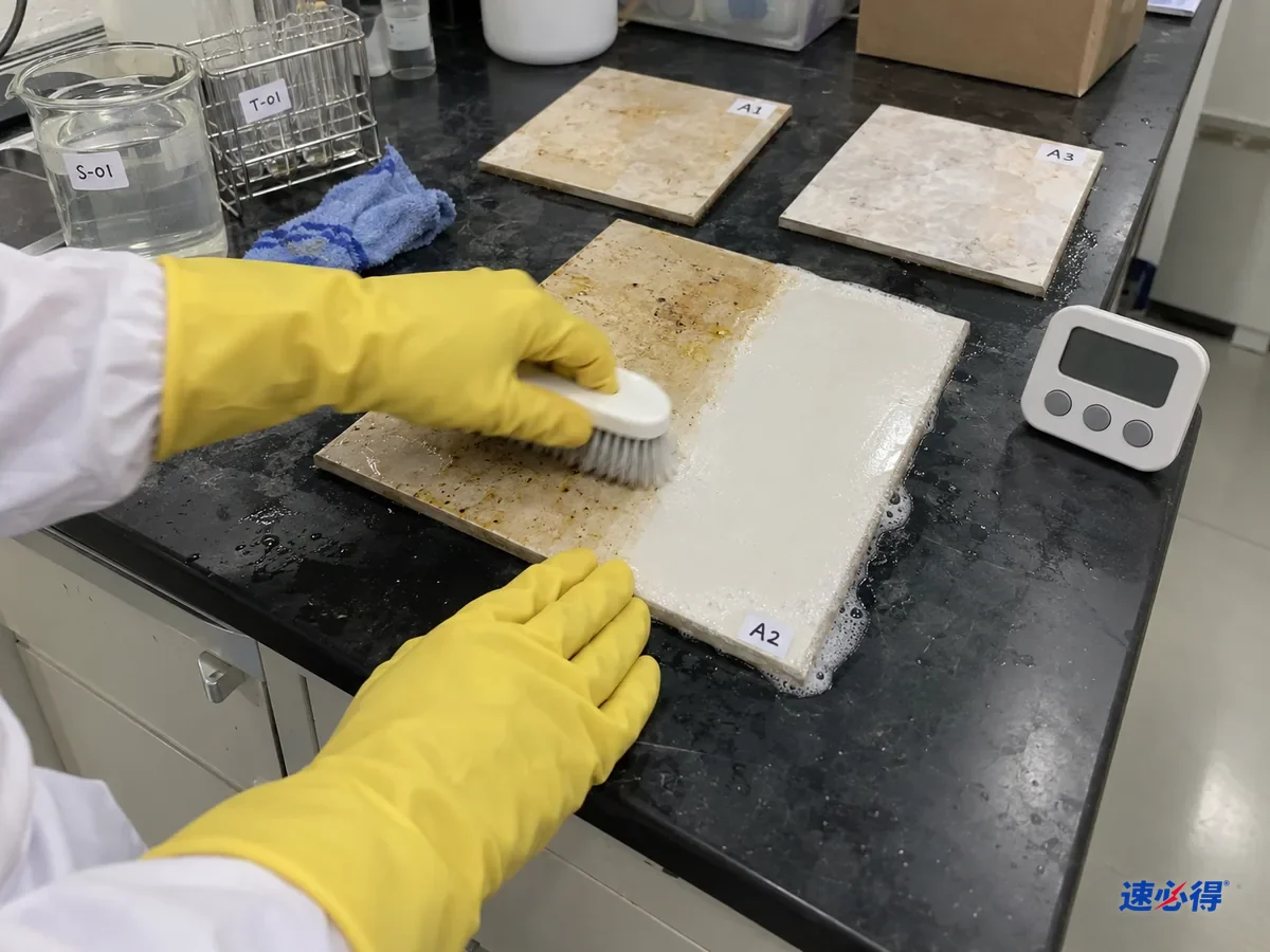实验台前，戴黄色橡胶手套的手和前臂用刷子刷瓷砖样板上的油污，刷过的一段变净，样板边缘有少量泡沫，计时器摆在旁边。
