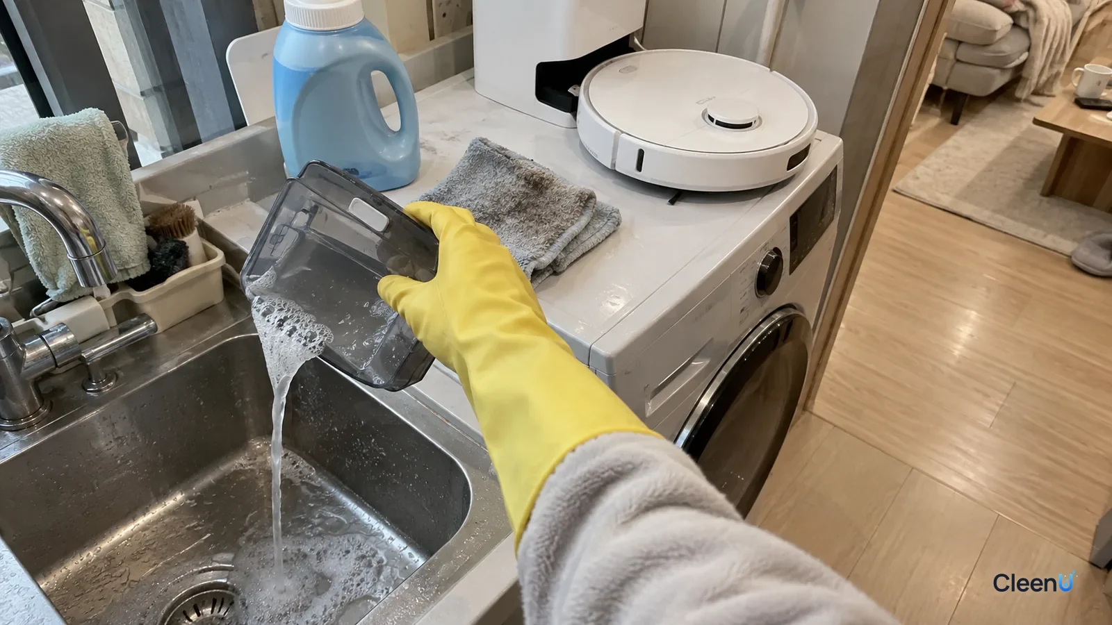 A yellow-gloved hand empties a robot mop's clean water tank into the laundry sink, a laundry detergent jug on the counter.
