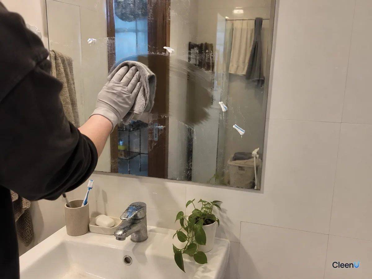 From a step back: a light gray nitrile-gloved hand wipes toothpaste spots from the mirror center outward, one area clean while a few white specks remain, the vanity below.