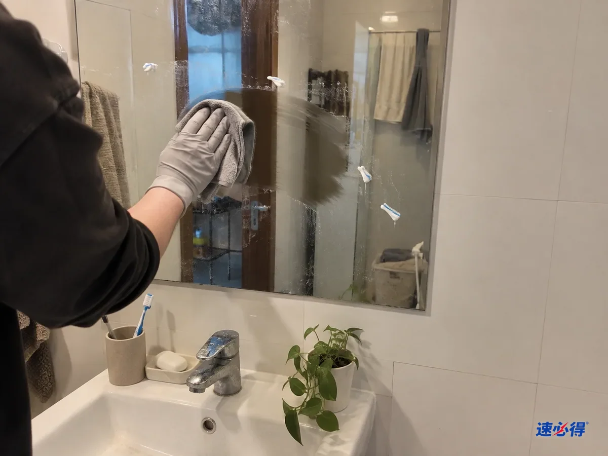 一步外看，戴浅灰色丁腈手套的手用超细纤维布从镜子中间往外擦牙膏点，擦过的一片干净，还有几点白色牙膏痕没擦到，下方是洗手台。