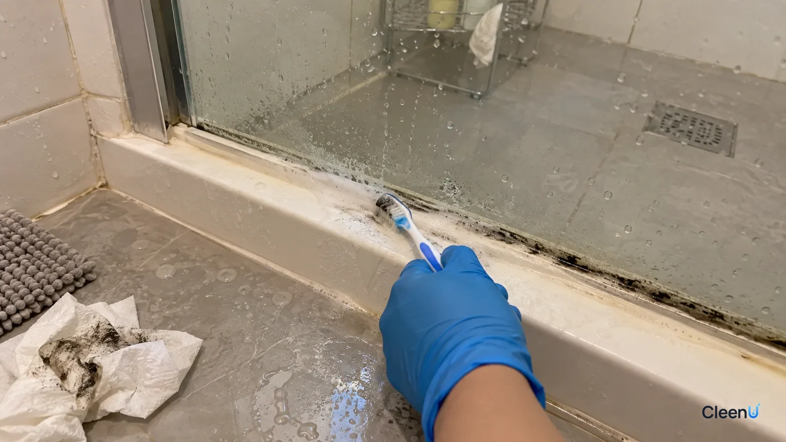 A gloved hand scrubs moldy shower caulk with an old toothbrush, one section lighter while the next still shows a black line, window open