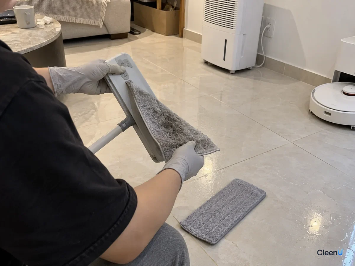 Person seen from behind in light grey nitrile gloves pulling a soaked microfiber pad off a flat mop, a dry pad on the floor, condensation beads still on the tiles