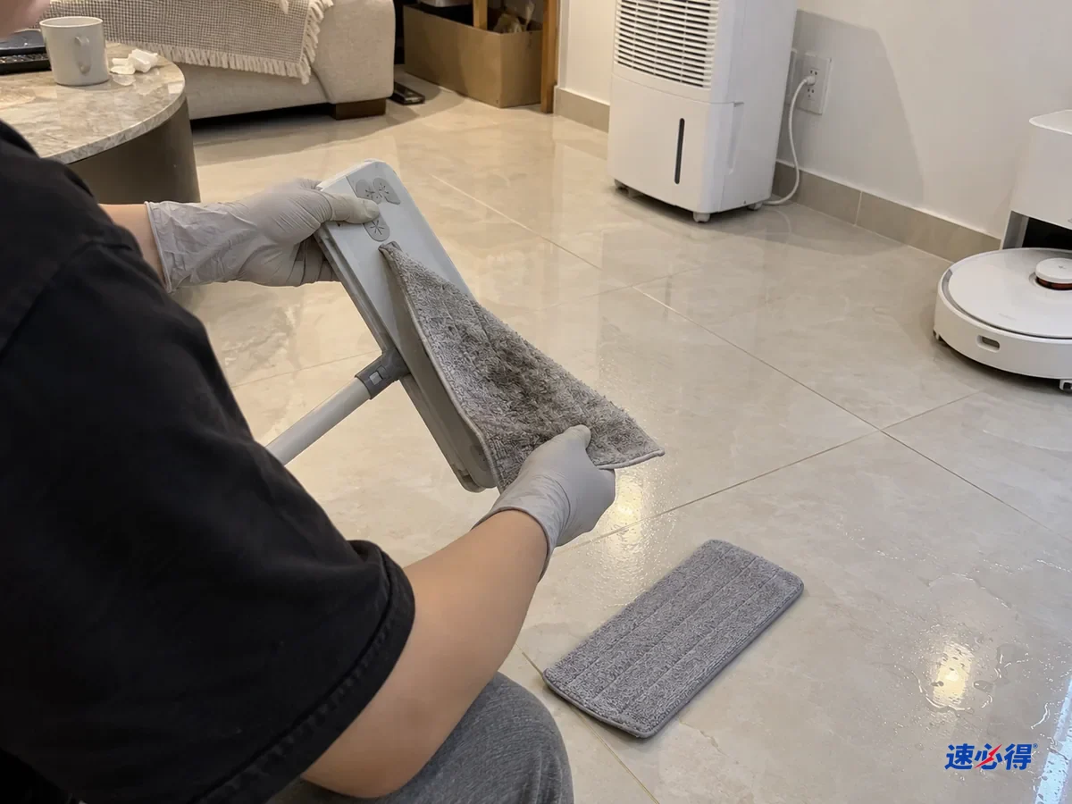 半身背影的人戴浅灰色丁腈手套，把一块吸饱水的超细纤维拖布从平板拖把上取下，地上放着一块干拖布，瓷砖上还有凝结水珠