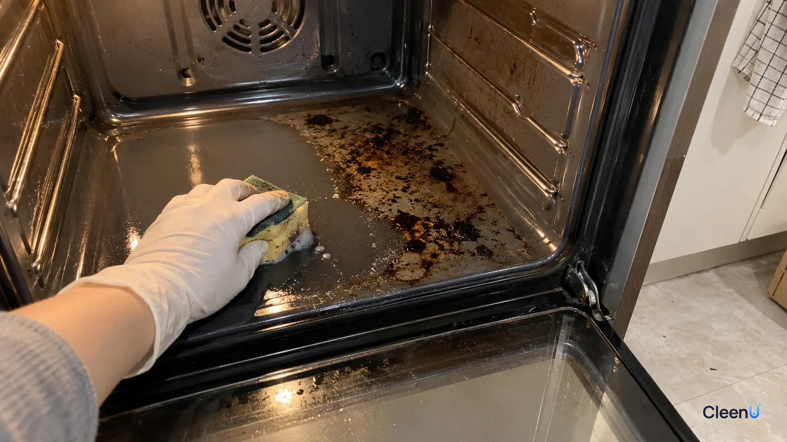 A gloved hand sponges the glossy enamel oven floor, one patch clean beside baked-on spots, the gray side liners left untouched